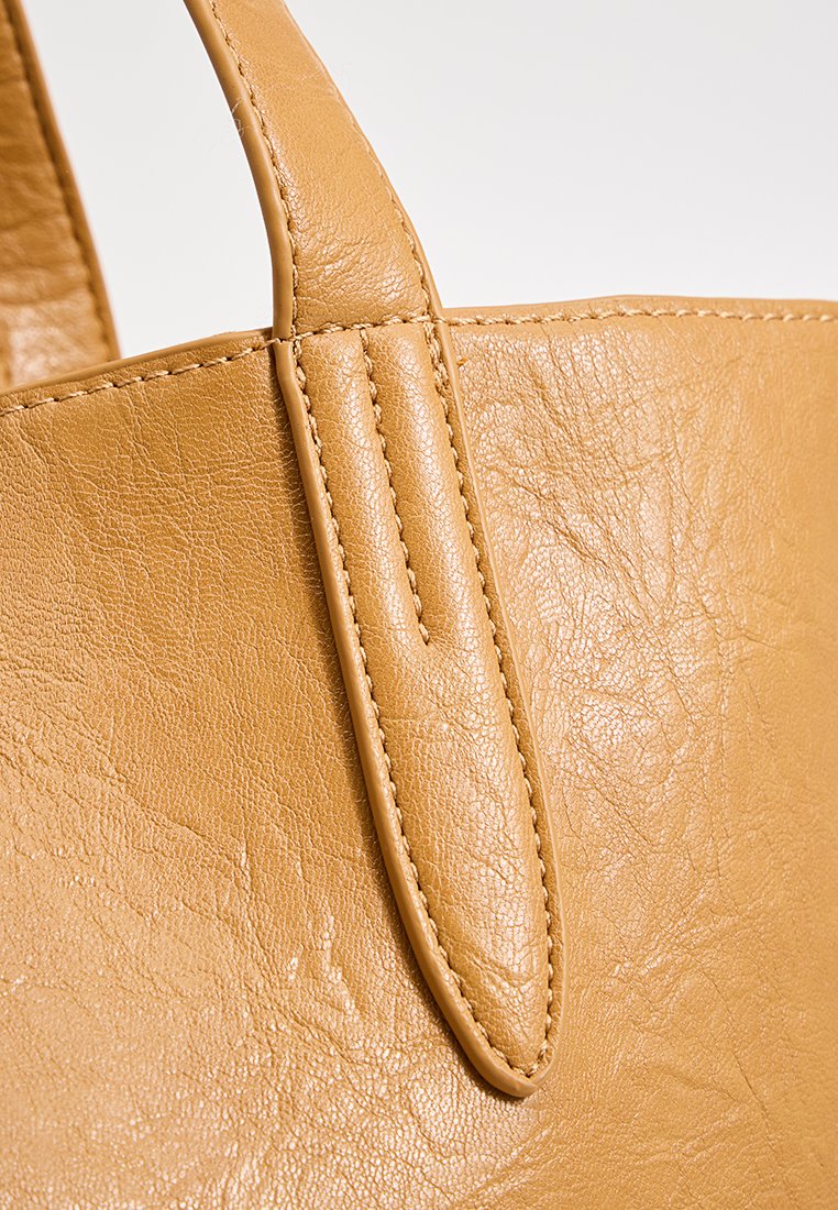 Détail d'un sac fourre-tout en cuir tan montrant la surface texturée, les coutures surpiquées et les poignées lisses, mettant en avant sa couleur et sa construction.