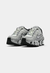 Para srebrno-czarnych sneakersów Nike Shox z widocznym sprężynowym amortyzatorem, białymi sznurówkami i czarnymi logo Nike swoosh na białym tle.