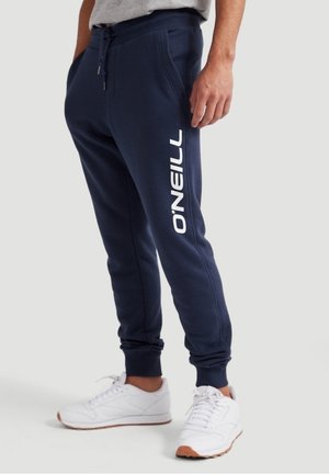 Homme portant un pantalon de jogging bleu marine avec le logo blanc O'NEILL sur la jambe et des baskets blanches devant un fond uni.