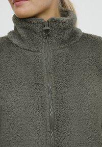 Groene fleece jas met hoge kraag, ritssluiting aan de voorkant en zachte, gestructureerde stof. Beschikt over een zilveren rits en subtiele stikdetails.