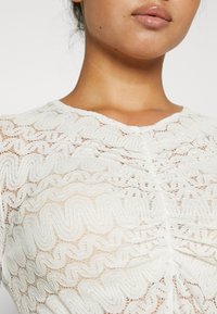 Gros plan sur une personne portant un haut en dentelle blanche avec des motifs brodés ondulés, montrant son cou, son menton et ses lèvres.