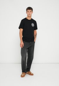 Camiseta de algodón negra con cuello redondo y estampado gráfico en el lado izquierdo del pecho. Combinada con jeans de mezclilla gris desgastados y zapatillas marrones con acentos beige.