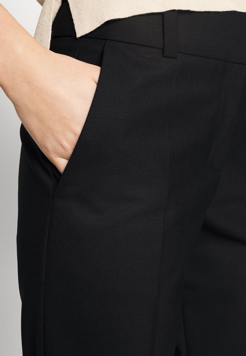 Hand in der vorderen Tasche von schwarzen maßgeschneiderten Hosen, mit einem beigefarbenen Oberteil, das teilweise darüber sichtbar ist.