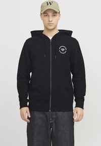 Sudadera negra con cremallera y bolsillo canguro, puños acanalados y dobladillo. Presenta un logo blanco en el lado izquierdo del pecho. Combinada con pantalones de mezclilla oscuros.