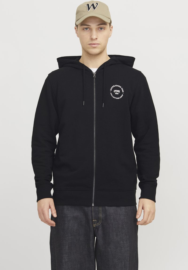 Sudadera negra con cremallera y bolsillo canguro, puños acanalados y dobladillo. Presenta un logo blanco en el lado izquierdo del pecho. Combinada con pantalones de mezclilla oscuros.