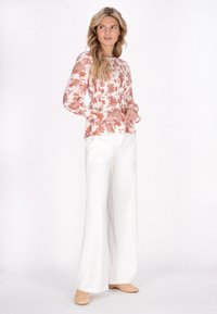 Blusa a maniche lunghe con motivo floreale in corallo e bianco, abbinata a pantaloni bianchi a vita alta e scarpe slip-on color beige chiaro.