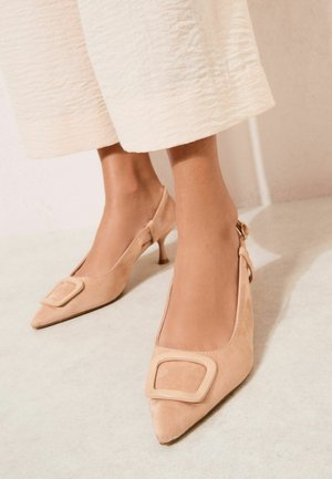 Kāju apavi ir gaiši brūni zamšādas augstpapēžu kurpes ar šauru degunu un liela kvadrātveida sprādzi, kopā ar krēmkrāsas teksturētām platām biksēm.