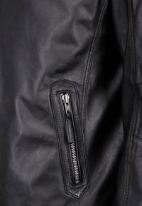 Nærbillede af en sort læderjakke med en lynlåslomme, glat overflade og detaljeret syning langs sømmene.