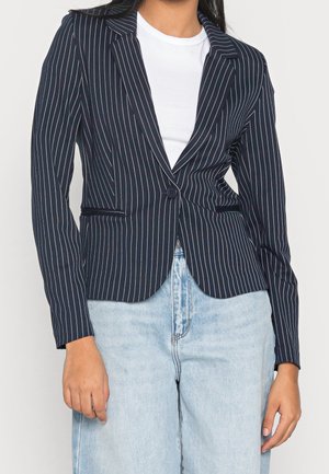 Frau trägt einen taillierten dunkelblauen Blazer mit weißen Nadelstreifen über einem weißen Hemd, kombiniert mit hellblauen, hoch taillierten Jeans.