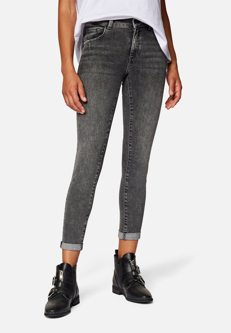 Femme portant un jean skinny noir délavé avec des revers, des bottines noires à boucles, et un haut blanc sur un fond uni.
