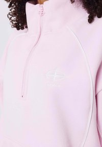 Lichtroze pullover van zacht materiaal, met een hoge kraag, een halfrits en een wit logo op de borst met contrasterende stiksels.