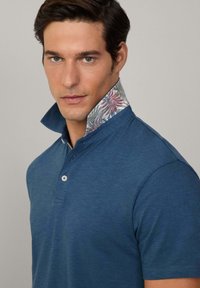 Blå polo skjorta med en texturerad yta, med ett blommigt mönster på den inre kragen och en treknapps placket.