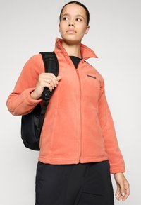 Veste en polaire corail avec une fermeture éclair intégrale, un col montant et un sac à dos noir. Logo de la marque en évidence sur la poitrine gauche. Tissu texturé.