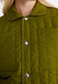Kviltad grön jacka med krage, tryckknappar och en bröstficka med en cirkulär metallaccent. Texturen verkar mjuk och vadderad.