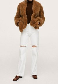 Brązowa kurtka z imitacji futra o puszystej fakturze, noszona na ciemnym golfie. Połączona z podartymi białymi dżinsami i brązowymi skórzanymi butami.