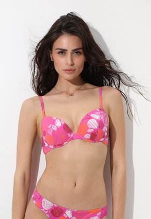 Donna con lunghi capelli scuri che indossa un bikini rosa con motivi floreali e conchiglie, in piedi davanti a uno sfondo bianco.