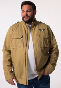 Giacca color khaki con colletto alto, due tasche sul petto e una toppa sulla spalla con un emblema alato. Indossata sopra una T-shirt bianca.