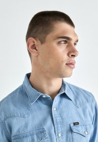 Jasnoniebieska denimowa koszula z klasycznym kołnierzykiem, dwiema kieszeniami na piersi oraz metalowymi guzikami. Posiada wkładkę z teksturowanej tkaniny i wzór w stylu western.