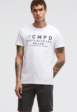 Mann mit Tattoos an Armen und Hals, trägt ein weißes T-Shirt mit dem Aufdruck "CMPD Contemporary Brand" und schwarze Shorts, vor einfachem Hintergrund.
