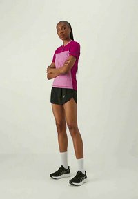 Frau steht mit verschränkten Armen, trägt ein pinkes und lila Sportshirt, schwarze Shorts, weiße Socken und schwarze Laufschuhe vor einem einfarbigen Hintergrund.