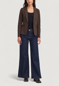 Blazer marrone sopra un top nero senza maniche, abbinato a jeans in denim blu scuro a gamba larga. Le caratteristiche includono rever sagomati e tasche laterali.
