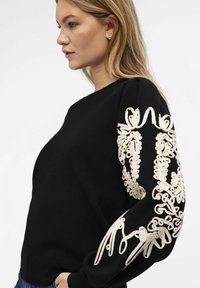 Pull noir avec broderie crème sur les manches, design à col rond, texture douce, silhouette ajustée et finitions côtelées.