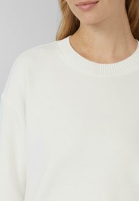 Vrouw in een witte geribde crewneck trui, met zichtbaar hals, schouders en een deel van haar gezicht, met een lichte huid en lichtbruin haar.