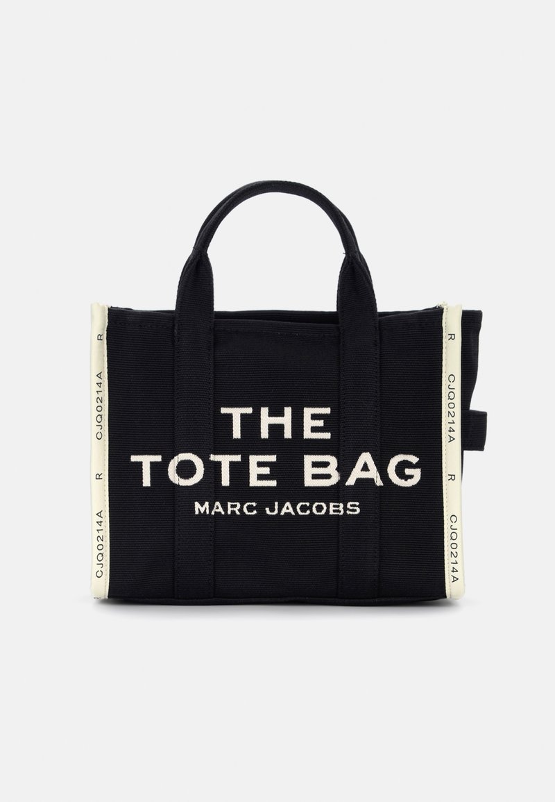 Mustat kangaskassit, joissa on valkoinen teksti, joka sanoo "THE TOTE BAG MARC JACOBS." Kassi on varustettu tukevilla kahvoilla ja minimalistisella muotoilulla.