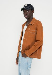 Veste en toile marron avec des coutures blanches et une poche poitrine. Portée sur un débardeur blanc, associée à un jean bleu clair et une casquette rayée.