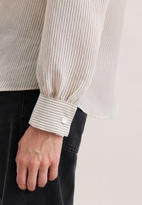 Nahaufnahme einer Hand, die seitlich ruht, bekleidet mit einem cremefarbenen Langarmshirt mit dünnen schwarzen vertikalen Streifen und schwarzen Hosen.
