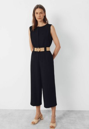 Kobieta stojąca przy białej ścianie, ubrana w czarny kombinezon bez rękawów z szerokimi, krótkimi nogawkami, beżowy pasek z okrągłą klamrą oraz beżowe sandały na słupku.