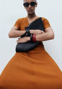 Black woven handbag held close with two circular bangles, one glossy red and one matte black, worn over an orange, short-sleeved dress.