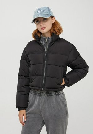 Giacca a vento nera con collo alto e chiusura con zip, caratterizzata da cuciture orizzontali e polsini elasticizzati. Abbinata a pantaloni grigi e un cappellino azzurro chiaro.