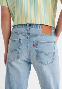 Personne portant un jean Levi's 501 bleu clair et une chemise rayée jaune, vue de dos à hauteur de la taille.