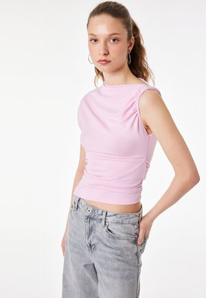 Jeune femme avec des boucles d'oreilles en anneau portant un haut rose pâle sans manches froncé et un jean gris ample sur un fond blanc uni.