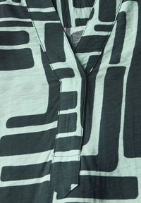 Korte mouwen shirt van mintgroene stof met gedurfde zwarte geometrische patronen, een V-hals en zichtbare stiksels langs de randen.
