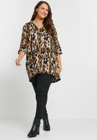 Tunika con stampa leopardata in marrone, nero e crema, con scollatura a V e maniche a tre quarti, abbinata a jeans skinny neri e stivali alla caviglia.