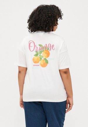 Une personne avec des cheveux bouclés porte un t-shirt blanc avec le texte "Orange" et des illustrations orange au dos, associé à un pantalon sombre.