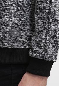 Felpa grigia e nera con texture, con polsini e orlo a costine. Presenta una patch rotonda con logo sul lato sinistro in basso.