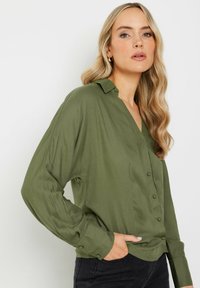 Blouse olive green à manches longues avec un col en V, fermetures à boutons et coupe décontractée. Texture lisse, avec des poignets roulés et un léger éclat.