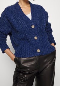 Mörkblå stickad kofta med V-krage, ribbad struktur och tre stora beige knappar, tillsammans med svarta läderbyxor.