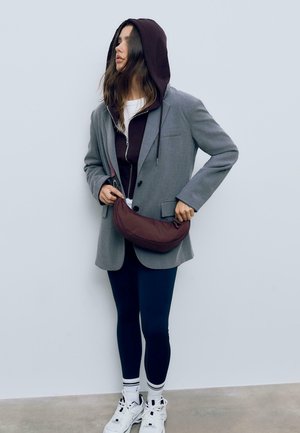 Grau blazer über einem lila Hoodie, kombiniert mit navy Leggings, weißen Sneakers und einer braunen Umhängetasche. Das Outfit zeichnet sich durch geschichtete Texturen aus.