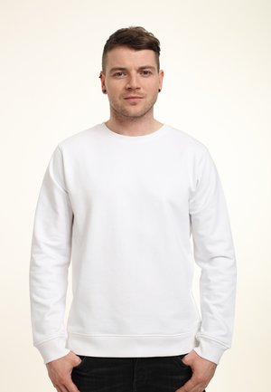 Jonge man met kort bruin haar en oorbellen, gekleed in een eenvoudige witte lange mouwen sweatshirt en zwarte broek, staand met zijn handen in zijn zakken.