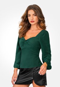 Blusa de color verde oscuro con una silueta ajustada, escote cuadrado y mangas largas abullonadas. Combinada con una mini falda de cuero negra con una abertura lateral.