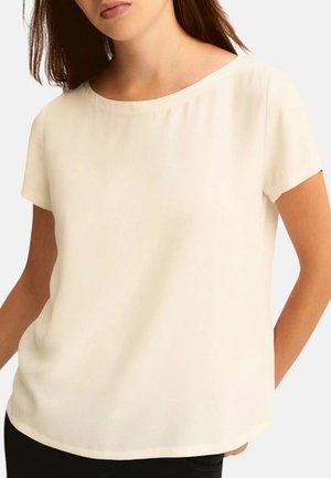 Femme portant un chemisier ivoire à manches courtes avec un col rond, vue des épaules aux hanches sur un fond uni.