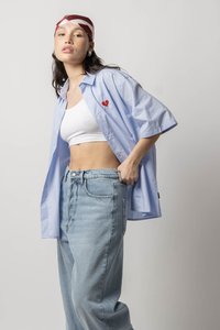 Lichtblauwe, korte mouwen overhemd met knopen en een rood hartapplicatie, gedragen over een witte crop top en gecombineerd met lichtblauwe hoge taille jeans.