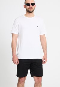 Man draagt een eenvoudige witte t-shirt, zwarte korte broek en zwarte zonnebril, staand tegen een witte achtergrond.