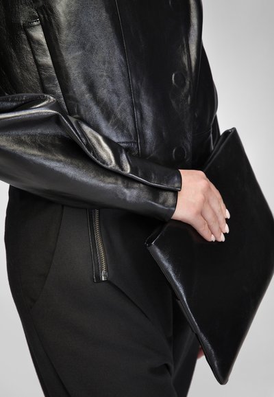 Čierny kožený clutch držaný v ruke, s hladkou textúrou a trojuholníkovým tvarom. Čiastočne je viditeľná priliehavá čierna bunda so lesklými rukávmi.