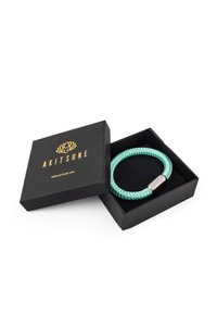 Bracciale intrecciato in turchese con chiusura metallica, contenuto in una scatola nera con accenti dorati e il nome del marchio "AKITSUNE."