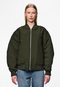Casaco bomber verde-azeitona com fecho de correr na frente, punhos canelados e corte oversized. Apresenta dois bolsos laterais. Usado sobre um denim casual.